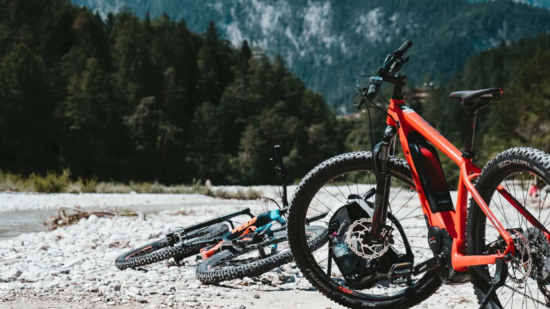 Zwei Mountainbikes auf einem Kiesufer, Wald und Berge im Hintergrund, sonniges Wetter.