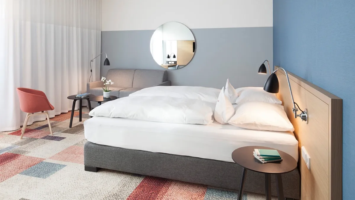 Modernes Hotelzimmer mit Doppelbett, Sofa, runder Spiegel, Nachttisch und farbigem Teppich. Neutrale Farbgebung und Lampe.