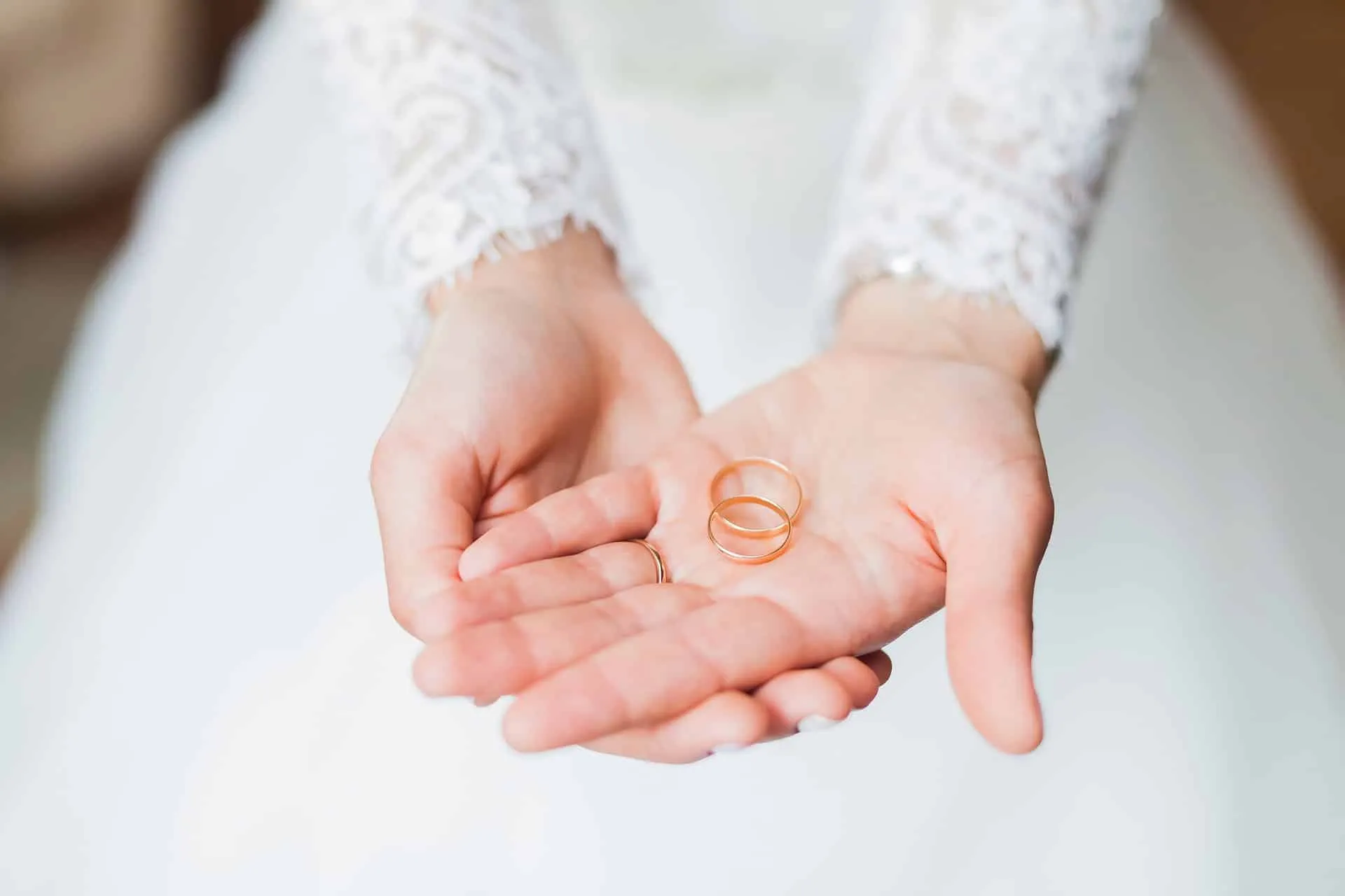 Hände in Spitzenkleid halten zwei goldene Eheringe, symbolisch für Hochzeit oder Verlobung.