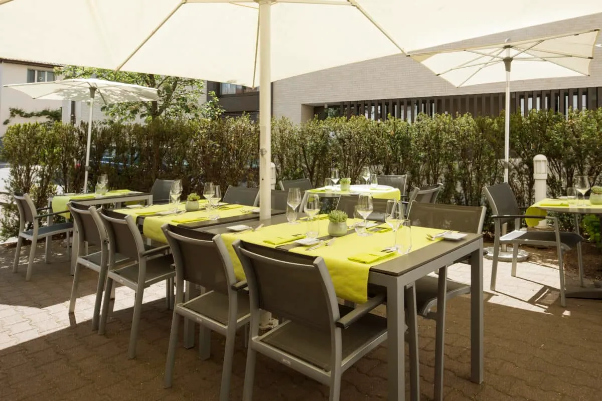 Sonniger Außenbereich eines Restaurants mit gedeckten Tischen, gelben Tischläufern, Sonnenschirmen und umgebender Hecke.