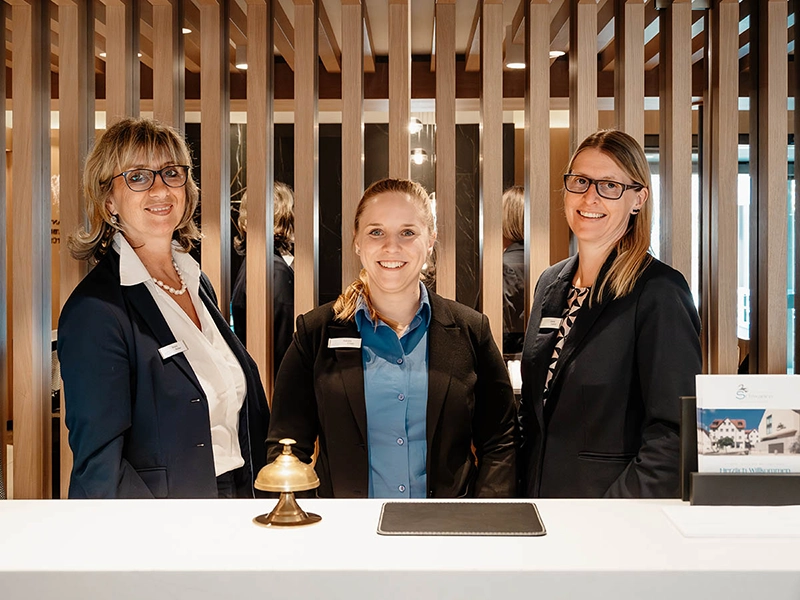Drei Frauen in Business-Outfits lächeln an einem Empfangstresen mit Glocke, im Hintergrund Holzgitterwand.