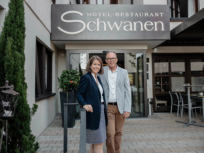 Paar vor dem Eingang des Hotels und Restaurants Schwanen, lächelnd, Geschäftskleidung, sommerlicher Tag, Außenbereich.