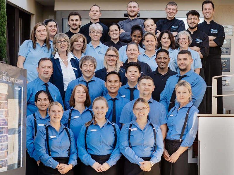 Gruppe von 28 Restaurantmitarbeitern in blauen Uniformhemden posiert lächelnd vor einem Gebäude.