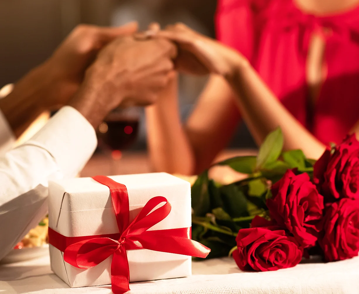 Paar hält Hände über Tisch mit Geschenk, roten Rosen und Glas Rotwein.