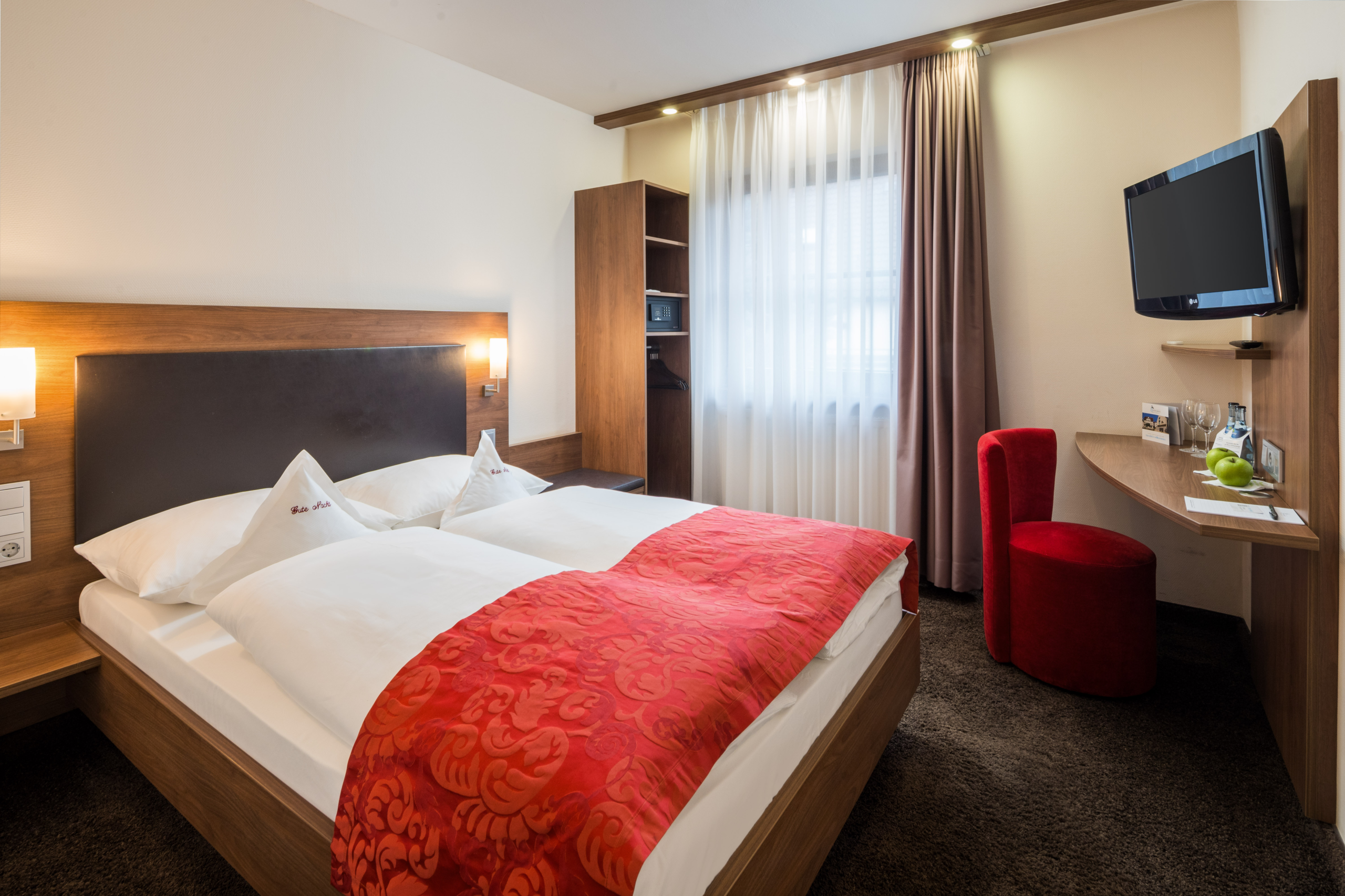 Hotel-Schwanen-Koengen_Queensize-Zimmer Helles Hotelzimmer mit Doppelbett, rotem Überwurf, TV, kleinem Schreibtisch, rotem Sessel und Fenstervorhängen.