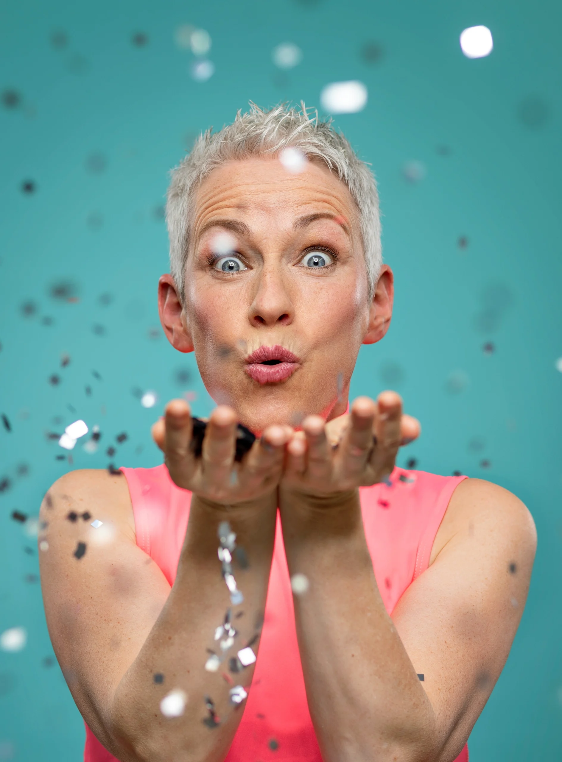 Frau mit kurzen Haaren pustet Konfetti aus ihren Händen in einem farbenfrohen Studio mit türkisfarbenem Hintergrund.