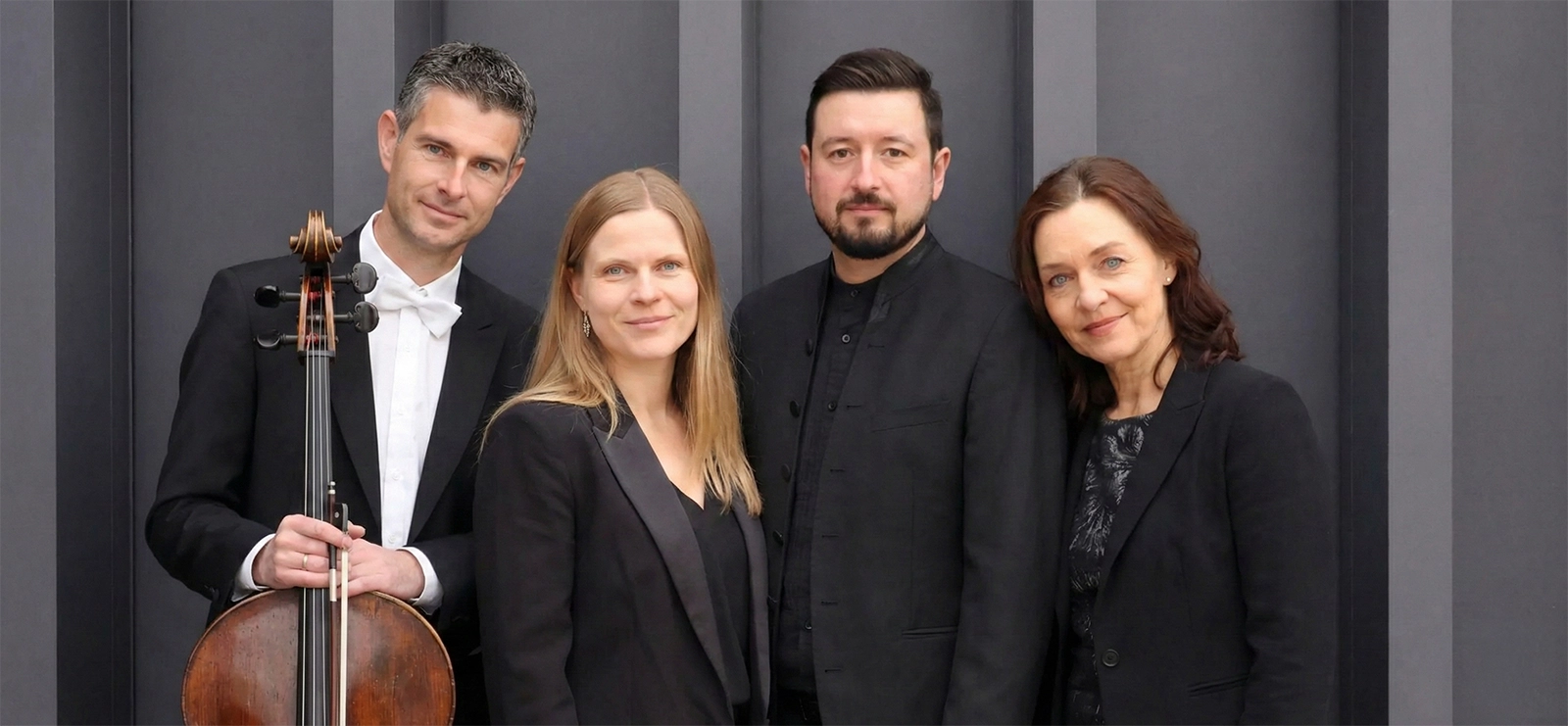 Vier Menschen in schwarzer Kleidung posieren vor einer grauen Wand, einer hält ein Cello.