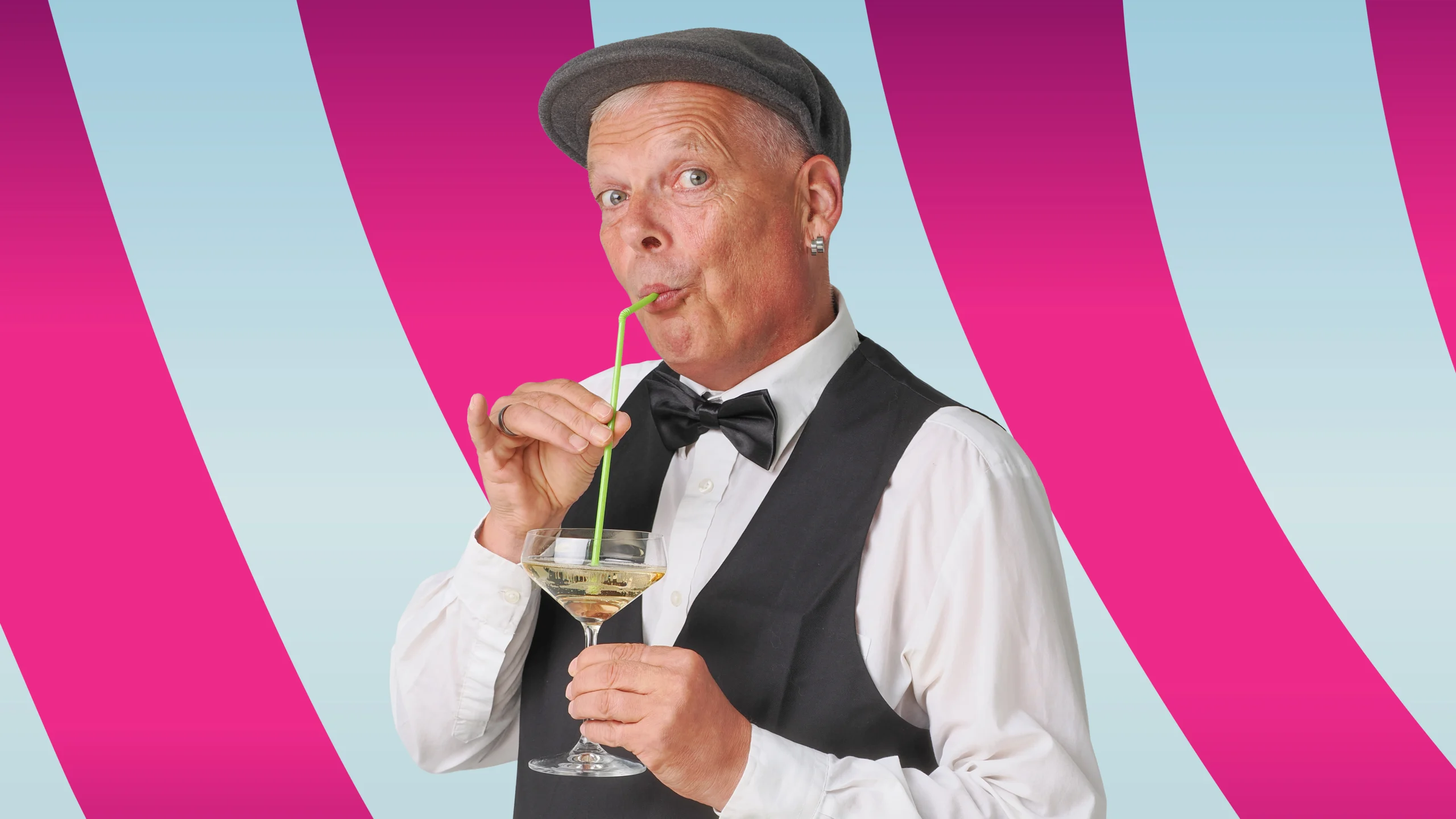 Mann mit Fliege und Schiebermütze trinkt aus einem Cocktailglas mit grünem Strohhalm vor gestreiftem Hintergrund.
