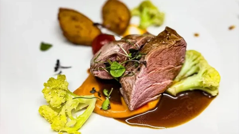 Schwanen-Koengen-Speisekarte-Restaurant-Unsere-Empfehlung-600×375 Gegrilltes Fleisch mit Kartoffeln, grünem Gemüse, Soße und Kräutern auf einem weißen Teller angerichtet.