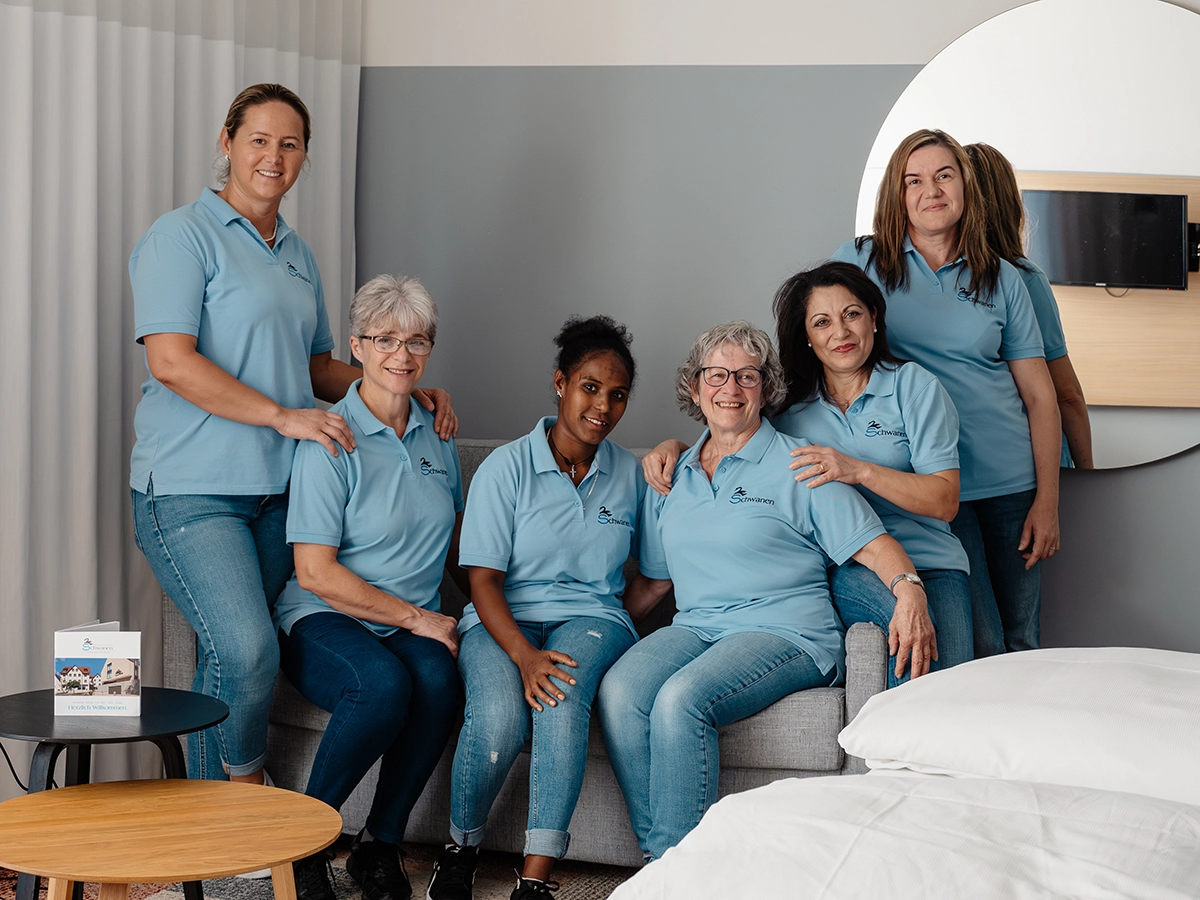 Sechs Frauen in blauen Poloshirts sitzen im modernen Raum auf einem Sofa, umgeben von weißen Bettwäsche und neutralen Farben.