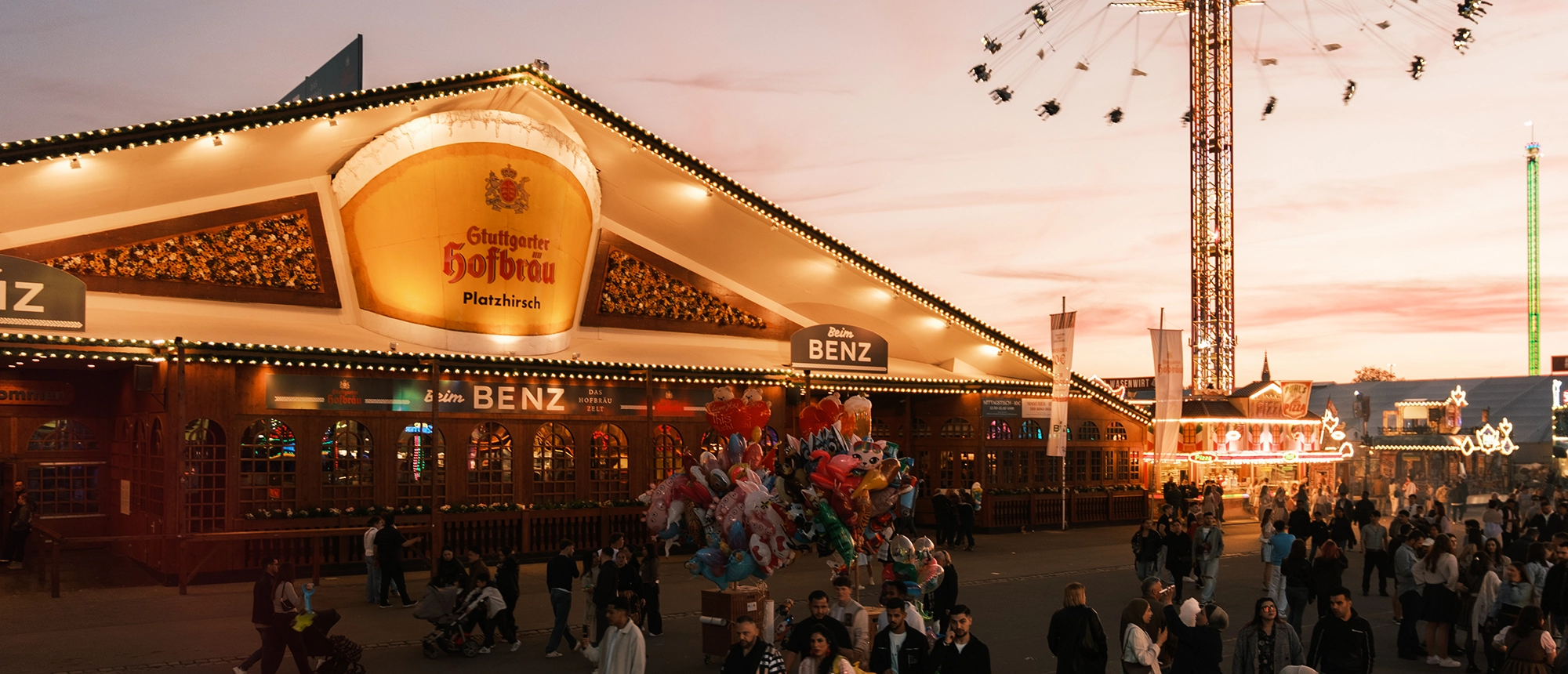 Menschen schlendern vor einem Festzelt mit Lichtern und einem großen Fahrgeschäft im Hintergrund, Abendstimmung.