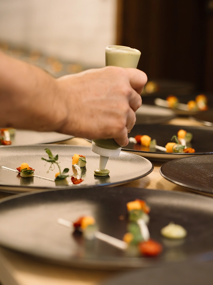 Schwanen_Food_Vorbereitung Koch spritzt grüne Sauce auf schwarze Teller, die mit buntem Gemüse und Kräutern dekoriert sind.