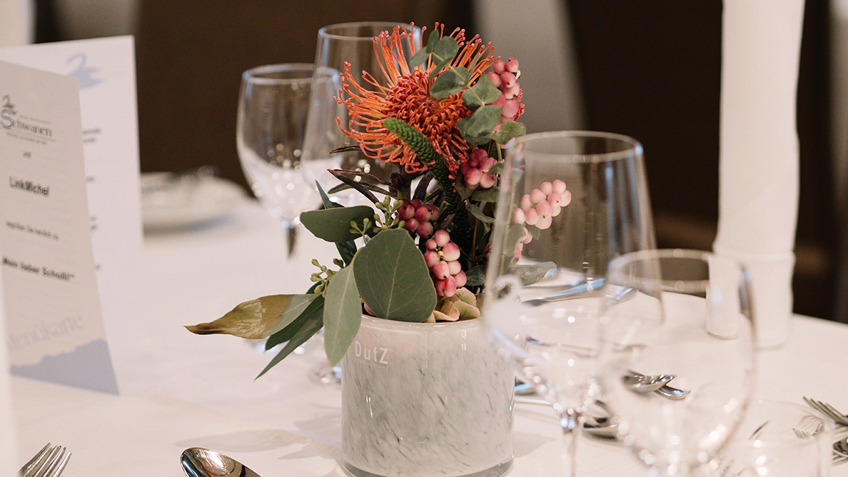 Schwanen_Kultur_Genuss_Deko Alter-Text: Blumenschmuck in einem weißen, marmorierten Topf, umgeben von Gläsern auf einem gedeckten Tisch.