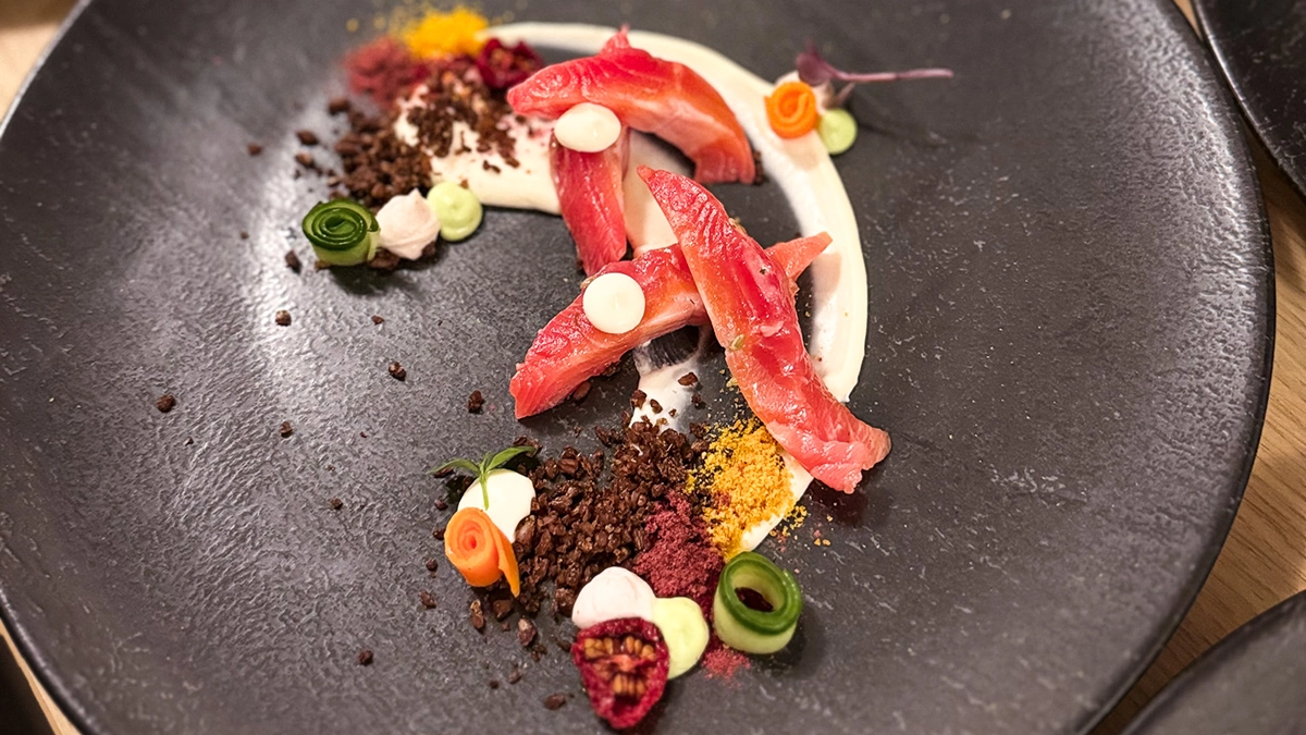 Schwanen_Kultur_Genuss_Essen Kunstvoll angerichtete Fischstücke auf dunklem Teller, umgeben von bunten Saucen und Gemüsestreifen.